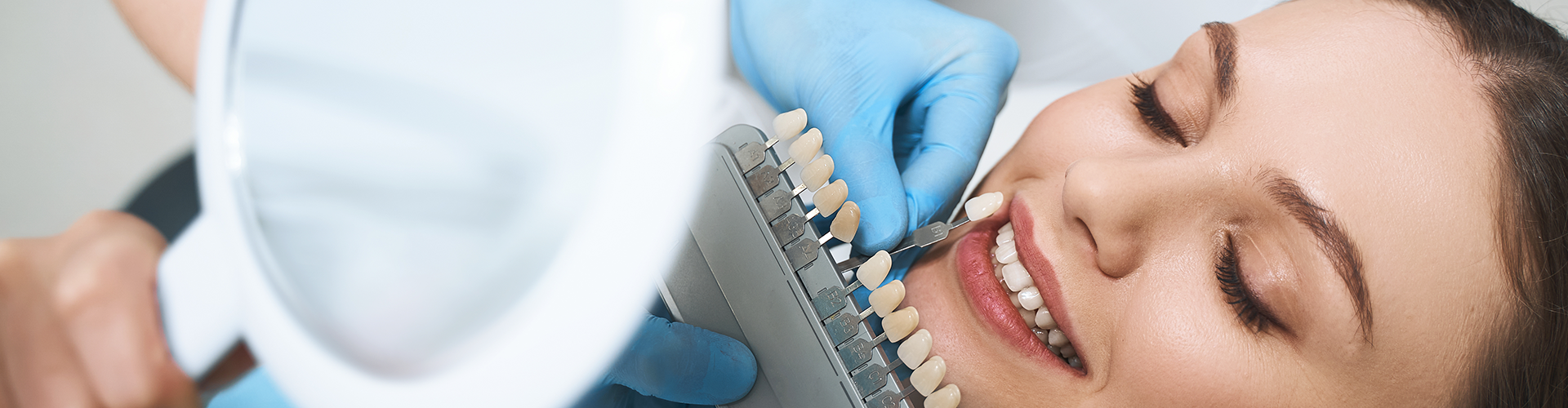 Gloved hands holding tooth shade-matching tool against woman's smile.